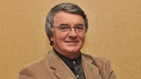 Portrét staršího muže s brýlemi v hnědém kabátě.
